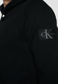 Černá mikina s kapucí vyrobená z měkkého materiálu, s texturovanou nášivkou s logem "CALVIN KLEIN JEANS" na levém rukávu a viditelnými švy.