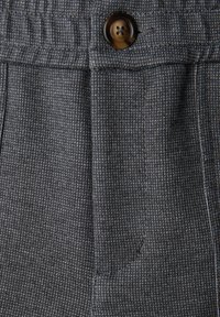 Tela gris con patrón que presenta un diseño de cuadros pequeños y texturizado. Hay un botón marrón cerca de un pliegue, con un corte casual y detalle en la cintura.