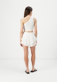 Fehér bordás crop top aszimmetrikus nyakkivágással, fehér, réteges szoknyával párosítva, amely rétegezett fodros dizájnnal rendelkezik. Fekete szandálok.