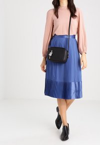 Black leather crossbody bag with a textured finish, featuring a silver charm accent and adjustable strap, paired with a pink blouse and blue pleated skirt.