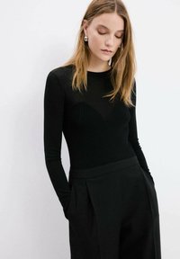 Mujer con cabello rubio liso, lleva pendientes de aro plateados, una blusa negra de manga larga acanalada y pantalones negros de talle alto con las manos en los bolsillos.