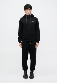 Νεαρός άνδρας στέκεται φορώντας μαύρο EA7 φούτερ με φερμουάρ και μπροστινές τσέπες, μαύρο ασορτί φόρμα και μαύρα αθλητικά παπούτσια, σε λευκό φόντο.