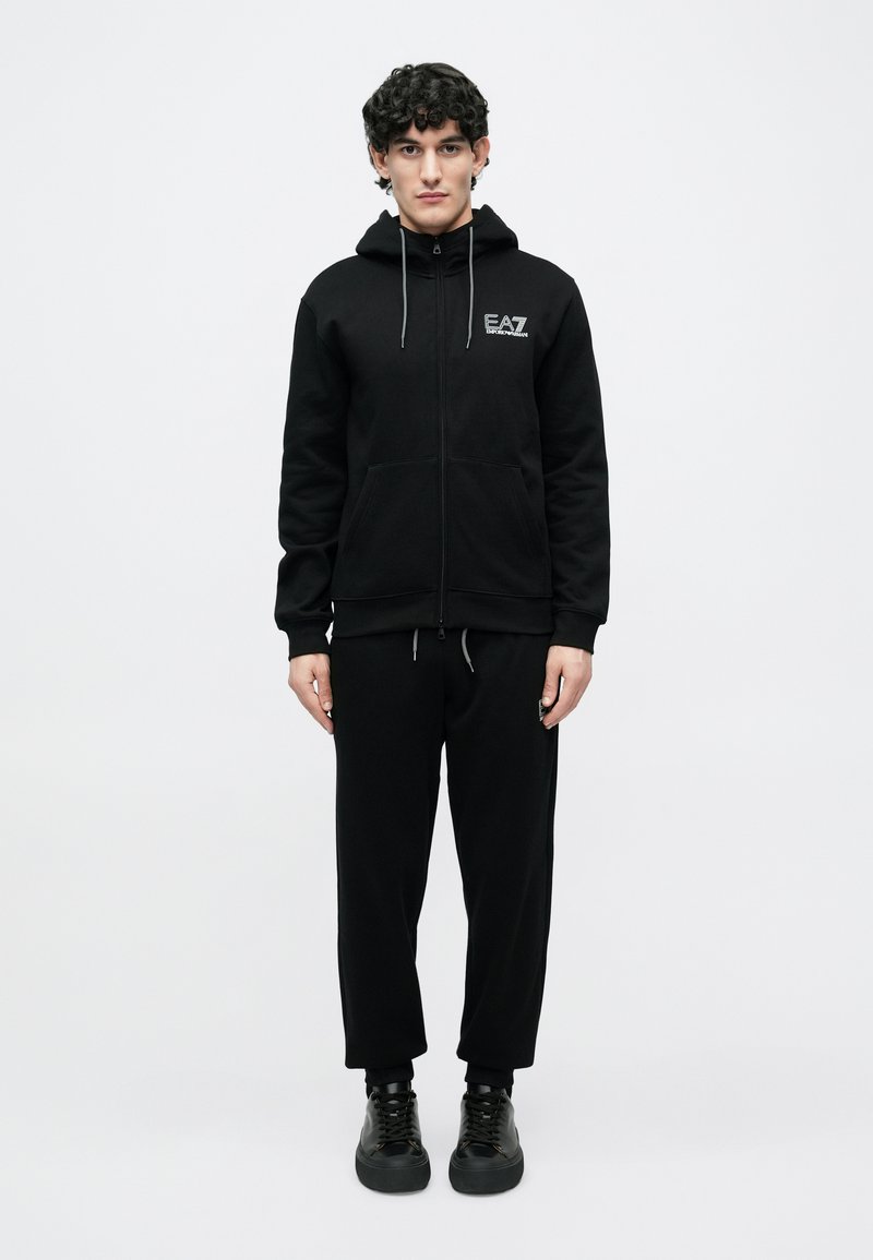 Νεαρός άνδρας στέκεται φορώντας μαύρο EA7 φούτερ με φερμουάρ και μπροστινές τσέπες, μαύρο ασορτί φόρμα και μαύρα αθλητικά παπούτσια, σε λευκό φόντο.