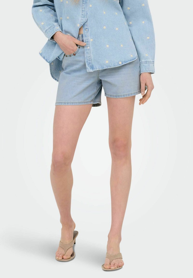 Femme portant un short en jean bleu clair et une chemise à manches longues assortie avec de petits imprimés marguerites, associée à des talons ouverts beiges.