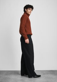 Donkerbruine ribgebreide turtleneck trui gecombineerd met zwarte wijde broek en spitse zwarte enkellaarzen. Eenvoudig, minimalistisch ontwerp.