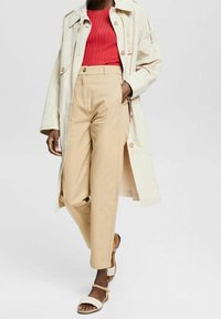 Lungo trench beige sopra un top rosso a coste, abbinato a pantaloni beige chiaro e sandali bianchi con cinturini. Texture morbide e vestibilità su misura.