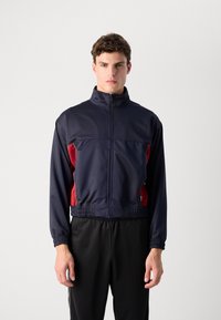 Ναυτική αδιάβροχη ζακέτα με κόκκινες πλευρικές λεπτομέρειες, πλήρης φερμουάρ, ψηλό γιακά, ελαστικούς μανσέτες και μέση; υφασμά από πολυεστέρα, λεία υφή.