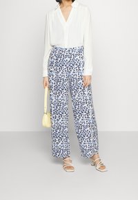 Woman wearing white blouse, blue and black leopard-print wide pants, white strappy heels, and holding a small yellow handbag.