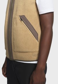 Beige Fleece-Weste mit weicher Textur, ausgestattet mit zwei diagonalen Reißverschlusstaschen und einem Reißverschluss in Kontrastfarbe. Akzentnähte am Saum.