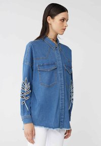 Camicia di jeans con colletto, due tasche sul petto e ricamo a motivo foglia bianca sulle maniche; presenta un orlo sfrangiato e una vestibilità rilassata.