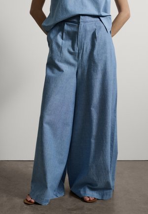 Persoon draagt lichtblauwe, hoge taille, wijde broek en een bijpassende top, staat met de handen achter de rug en voeten in sandalen.