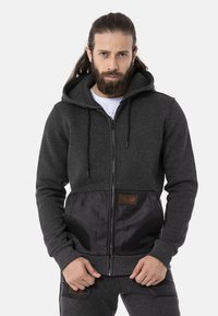Dark grey hoodie with a zipper, featuring a contrasting black nylon front panel and adjustable drawstring hood. Includes a logo patch.