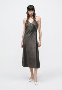 Tumši brūnas krāsas bezroku kleita ar vieglu krokainu faktūru, V veida izgriezumu un gaisīgu A līnijas siluetu. Kombinēta ar baltām augstpapēžu muleēm.