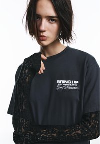 Črna majica s kratkimi rokavi z belim napisom. Dolgi črni čipkasti rokavi se raztezajo iz majice, kar v oblikovanje dodaja kontrast plasti teksture.