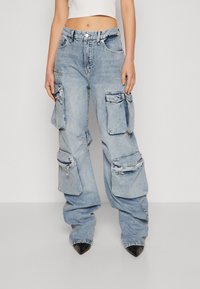 Žena ve světle modrých volných denimech cargo s několika velkými kapsami a černými špičatými botami, naznačující bílý zkrácený top.