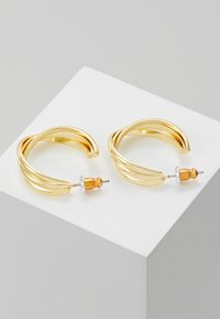Boucles d'oreilles en forme de créoles dorées avec un design tordu, arborant un accent argenté et un support en bois. Texture métallique lisse, forme circulaire.