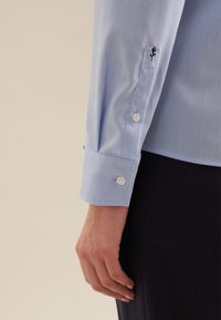 Lichtblauwe shirt met lange mouwen, voorzien van een op maat gemaakte manchet, witte knopen en een klein geborduurd rozenmotief op de manchet.