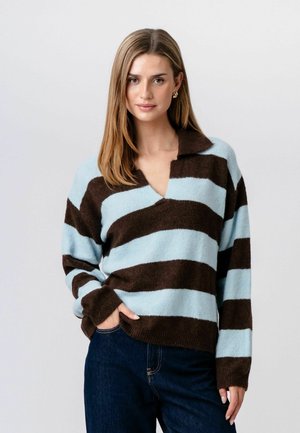 Woman with straight light brown hair wearing gold earrings, a blue and brown striped sweater with collar, and dark blue jeans, posing with hand in pocket.