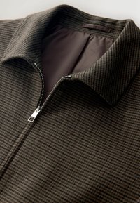 Texturierte, dunkelbraune Zip-Jacke mit feinen vertikalen Streifen. Sie hat einen spitzen Kragen und einen Metallschließe. Das Innenfutter ist einfarbig braun.