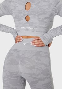 Grau camoufliertes Athleisure-Set mit einem Langarm-Oberteil, das drei runde Ausschnitte aufweist, und hochgeschnittenen Leggings; beide aus einem strukturierten Stoff gefertigt.