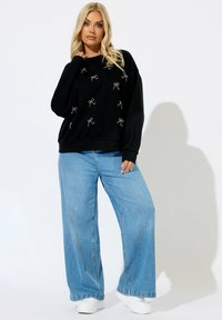 Sudadera negra con patrones de lazo bordados, corte relajado y puños acanalados. Combinada con jeans de pierna ancha de color azul claro y zapatillas blancas.