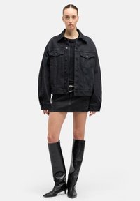 Kobieta ubrana w czarną, oversize'ową kurtkę dżinsową, czarną mini spódniczkę z ozdobnymi ćwiekami, czarny top, pasek i czarne skórzane kozaki sięgające do kolan.