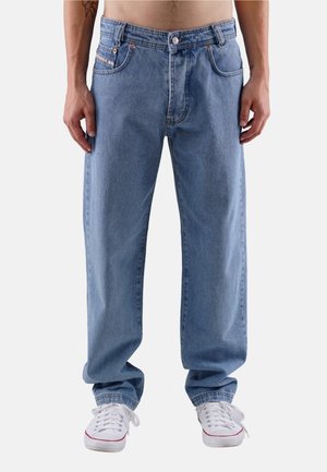 Man wearing loose-fitting blue jeans and white sneakers, standing with hands relaxed by sides against a plain background.