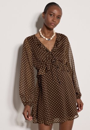 Donna che indossa un vestito marrone trasparente a pois con maniche lunghe e una collana bianca di perline, in piedi davanti a uno sfondo chiaro e semplice.