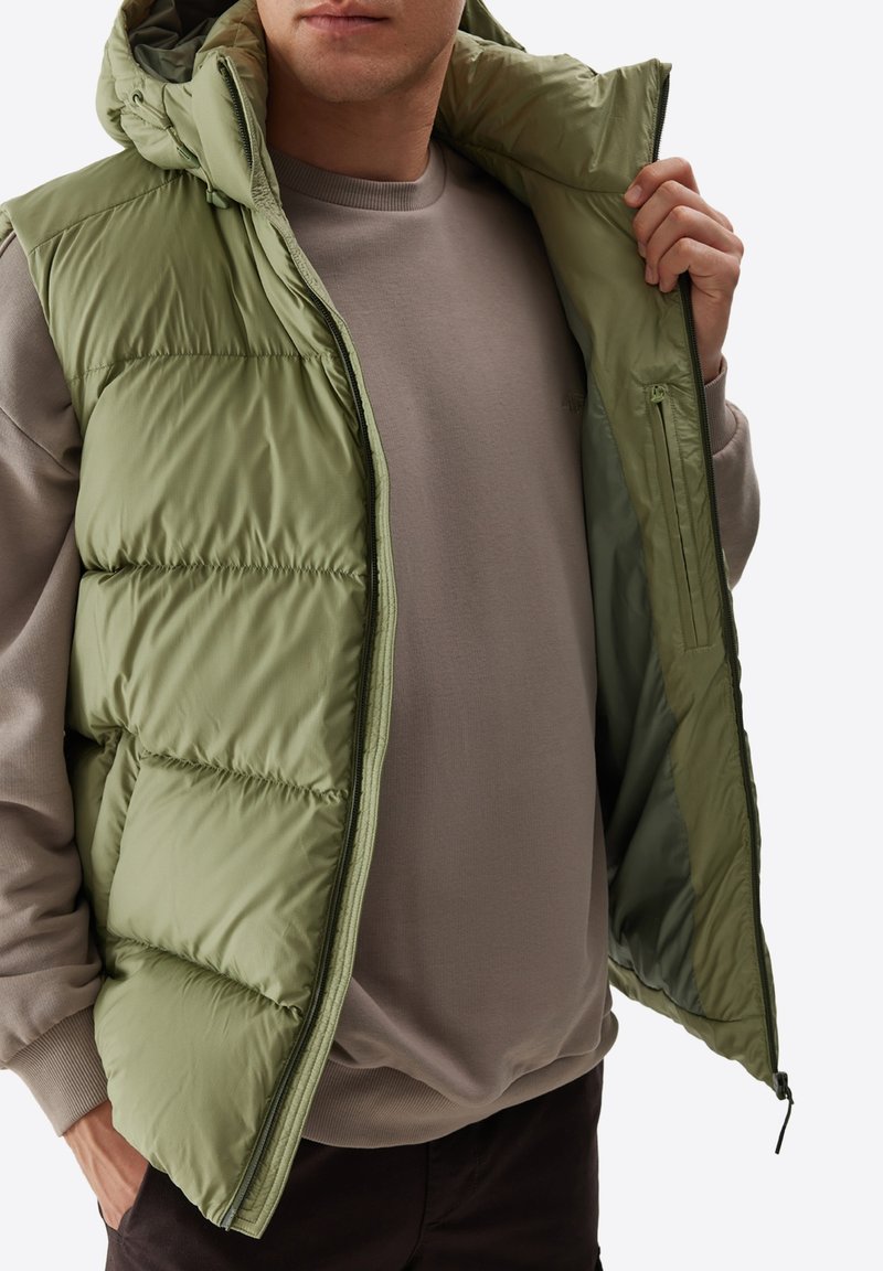 Oliwkowa kamizelka puchowa z kapturem, z pikowaną powierzchnią, z zamkiem z przodu i wewnętrzną kieszenią. Nosi się ją na szarym swetrze.