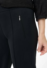 Zwarte op maat gemaakte broek met een zachte textuur, voorzien van een zijzak met ritssluiting en subtiele plooien. De tailleband is elastisch voor extra comfort.