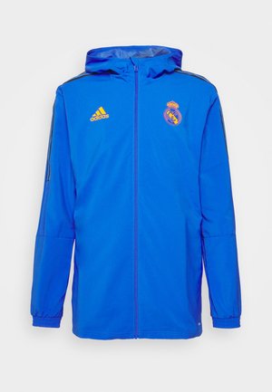 Blauer Adidas Sportjacke mit Reißverschluss und Kapuze, elastischen Bündchen, schwarzen Streifen an den Ärmeln sowie Real Madrid- und Adidas-Logos auf der Brust.