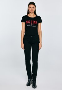 Nő fekete „BIG STAR” feliratú pólóban, fekete farmernadrágban és fekete bokacsizmában, világos szürke, egyszínű háttér előtt áll.