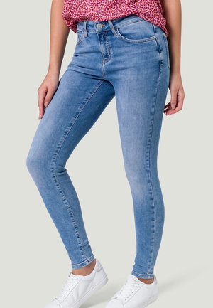 Femme portant un jean skinny bleu clair, un haut rose à motifs et des baskets blanches, debout les mains détendues le long du corps.
