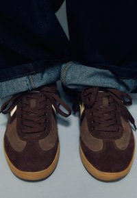 Hnědé semišové tenisky s hladkým koženým detailem, tmavě hnědými tkaničkami a podrážkou z gumové pryže; džíny s ohrnutými lemy, odhalující texturu denimu.