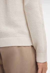 Helle beige Strickpullover mit einem strukturierten, offenen Muster, geripptem Saum und Ärmeln, kombiniert mit glattem, hellbraunem Stoff darunter.