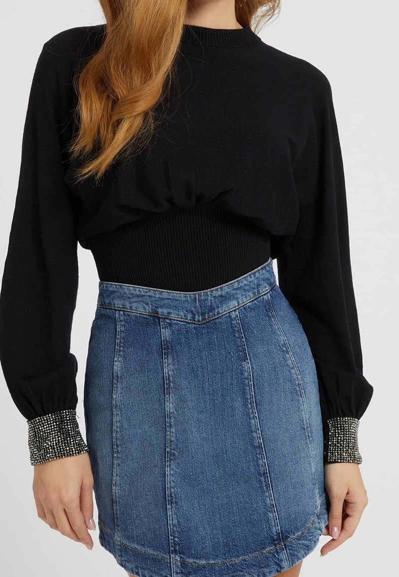 Femme portant un pull noir à manches longues avec des poignets ornés de strass et une jupe en jean bleue taille haute, debout devant un fond uni.