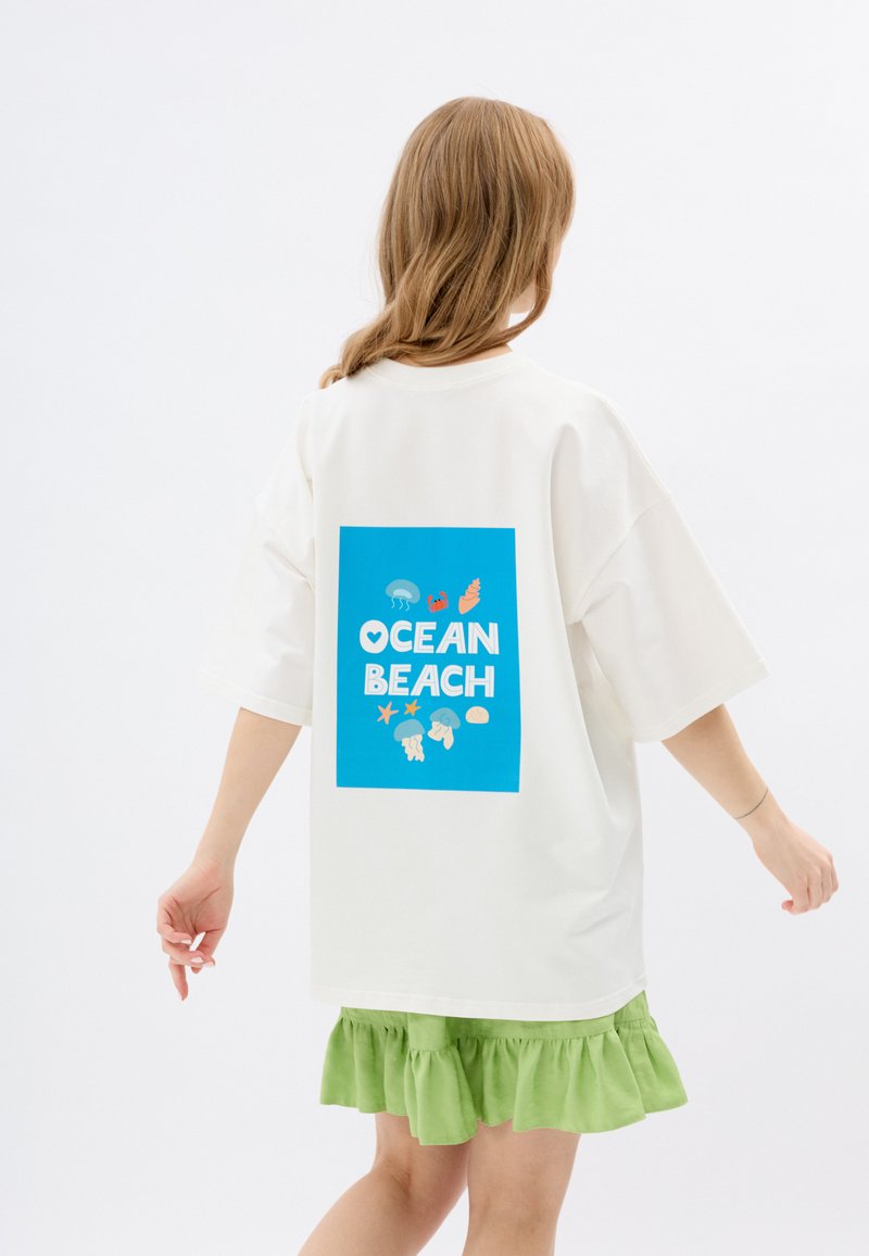 Przesadna biała koszulka z niebieską grafiką na plecach, przedstawiającą napis "OCEAN BEACH" oraz ikony związane z morzem. Posiada luźny krój i krótkie rękawy.