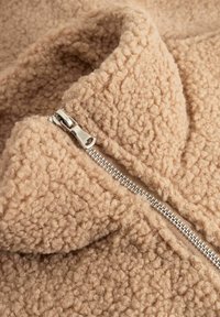 Bež flis jakna s teksturiranom površinom, ima srebrni patentni zatvarač i visok ovratnik. Tkanina izgleda mekano i toplo.