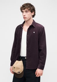 Σκούρα μωβ πουκάμισο από κορντόρι με κουμπιά στο εμπρόσθιο μέρος, λευκό εσώρουχο και μαύρο παντελόνι. Καφέ καπέλο μπέιζμπολ κρατημένο στο χέρι με μπλε έμβλημα.