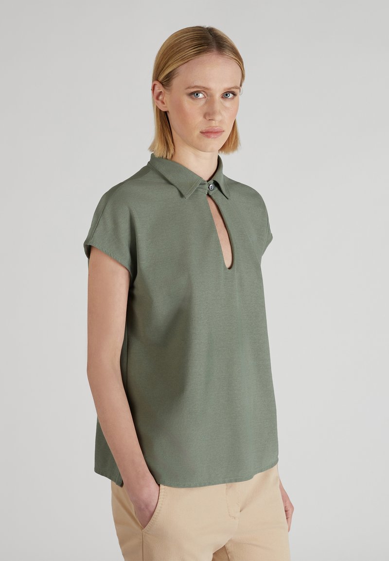 Kobieta z krótkimi blond włosami nosząca oliwkowozieloną bluzkę z krótkim rękawem, z kołnierzykiem i dekoltem w kształcie klucza, beżowe spodnie, stojąca na jednolitym tle.