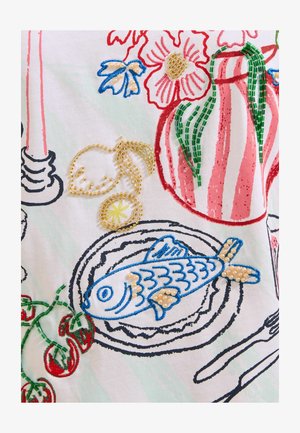 Beaded and embroidered fish on plate with lemon slices, tomatoes, and striped vase with flowers on patterned fabric.