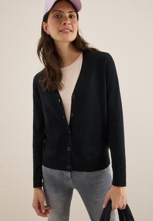 Zwarte cardigan met een v-hals, vijf knopen en een geribbelde zoom. Draagt over een lichtbeige top, gecombineerd met grijze jeans. Soepele textuur.