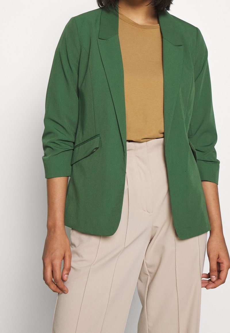 Personne portant un blazer vert sur une chemise marron et un pantalon beige taille haute, debout devant un fond clair uni.