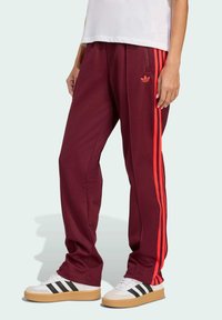 Burgunderfarbene Sporthose mit roten Seitenstreifen, hergestellt aus strukturiertem Stoff. Verfügt über ein gesticktes Logo an der linken Hüfte und Reißverschlusstaschen.
