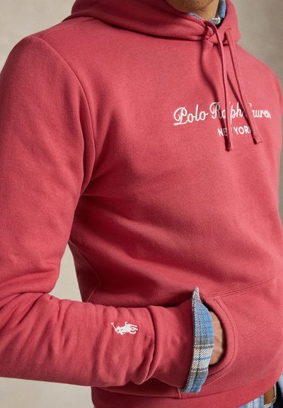 Άτομο που φοράει κόκκινο φούτερ Polo Ralph Lauren με λευκό λογότυπο και χέρι στην μπροστινή τσέπη πάνω από μανίκι μπλε καρό πουκαμίσου.