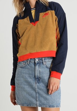 Femme portant un pull Nike marron et bleu marine avec des touches rouges et une jupe en jean bleu clair sur un fond clair uni.