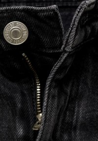 Acercamiento de una chaqueta de mezclilla negra que presenta un botón a presión de metal plateado y un zipper con dientes visibles. Tejido texturizado con detalles de costura.