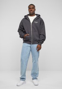 Charcoal zip-up hoodie with front pockets, ribbed cuffs, and a white graphic, paired with light blue straight-leg jeans and white sneakers.