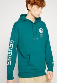 Tyrkysová mikina s kapucňou a predným vreckom, s šnúrkami na kapucni a bielym textom na rukávoch. Obsahuje grafické logo na hrudi. Mäkko a štruktúrovane tkanina.