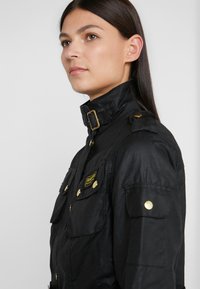 Black jacket featuring a high collar, gold hardware, shoulder epaulets, and multiple front pockets with a logo patch on the left side.